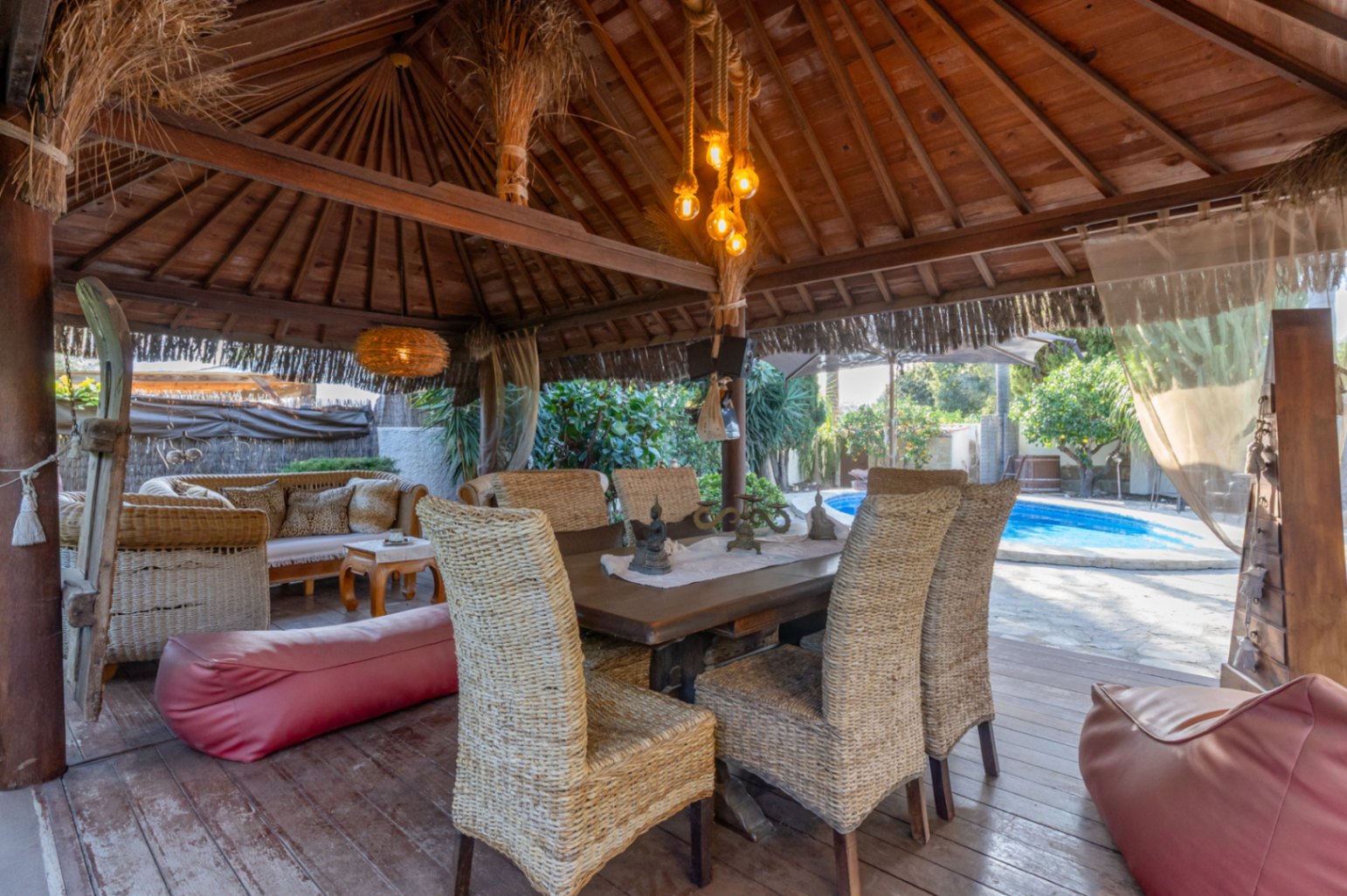 Pérgola de madera de estilo balinés con comedor exterior, sillas de mimbre y zona de estar junto a la piscina privada.
