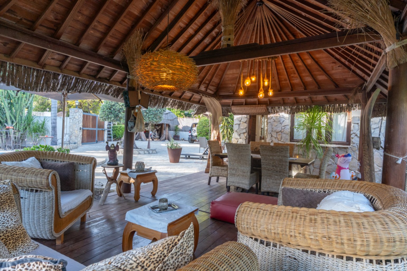 Pérgola de madera de estilo balinés con techos de vigas expuestas, zona de estar con sofás de mimbre y comedor exterior sobre suelo de tarima.