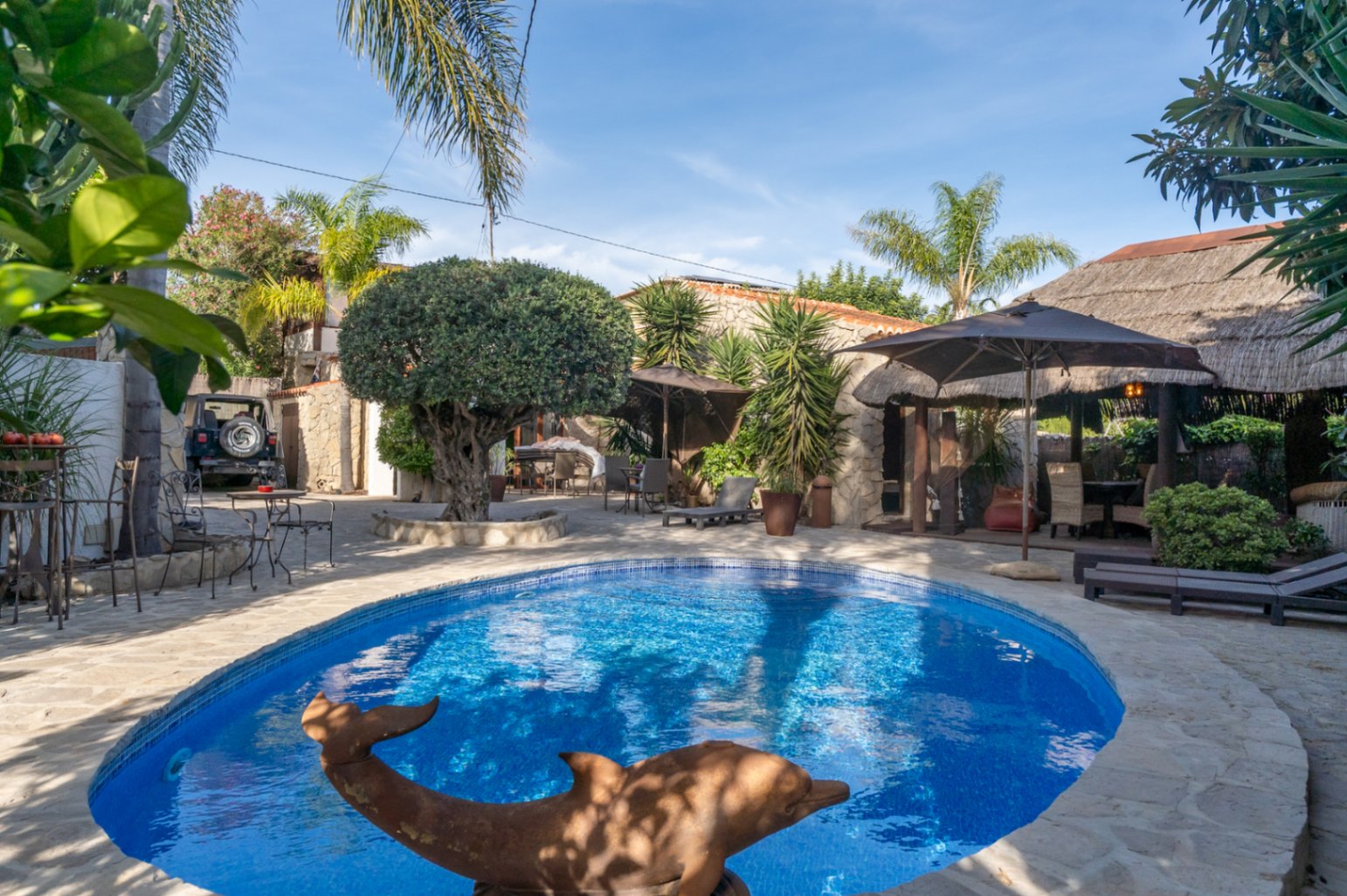 Piscina ovalada rodeada de pavimento de piedra natural, con una pérgola de estilo balinés, vegetación mediterránea madura y zonas de descanso.