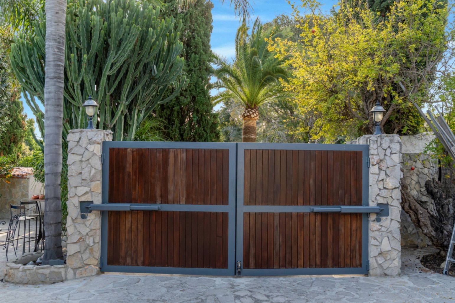 Puerta de acceso automática con paneles de madera y marco metálico, flanqueada por pilares de piedra natural y exuberante vegetación mediterránea.