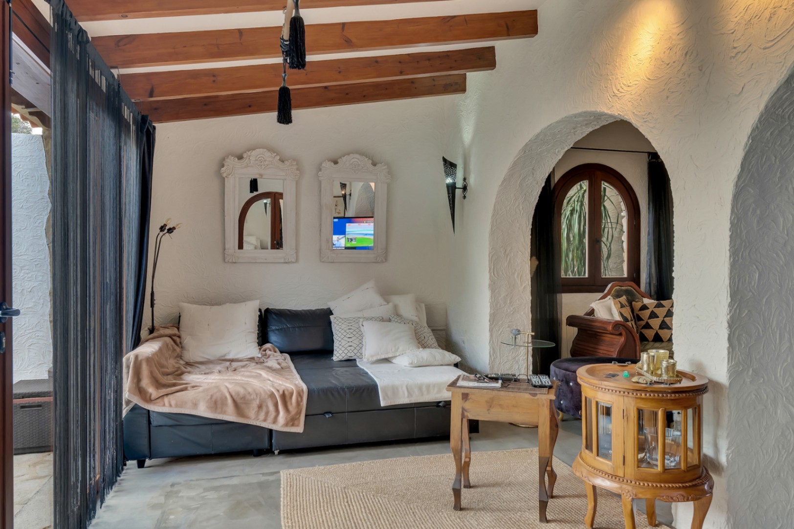 Salón acogedor con vigas de madera a la vista, paredes de yeso texturizado y arcos tradicionales. Incluye mobiliario de madera, espejos ornamentados y abundante luz natural.