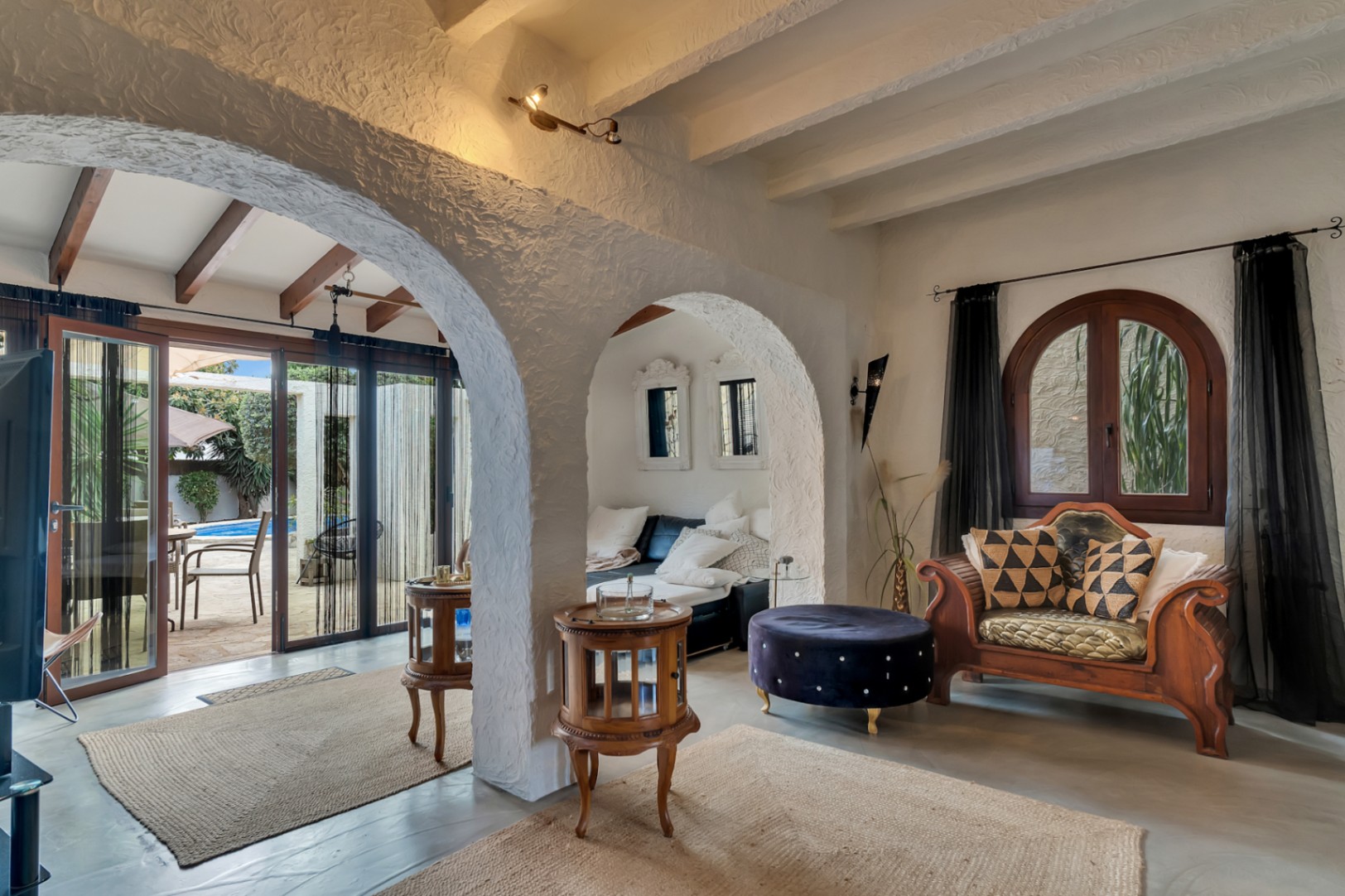 Salón con arcos de estilo mediterráneo, vigas vistas blancas y acceso directo a una naya acristalada con vistas a la piscina y jardín.