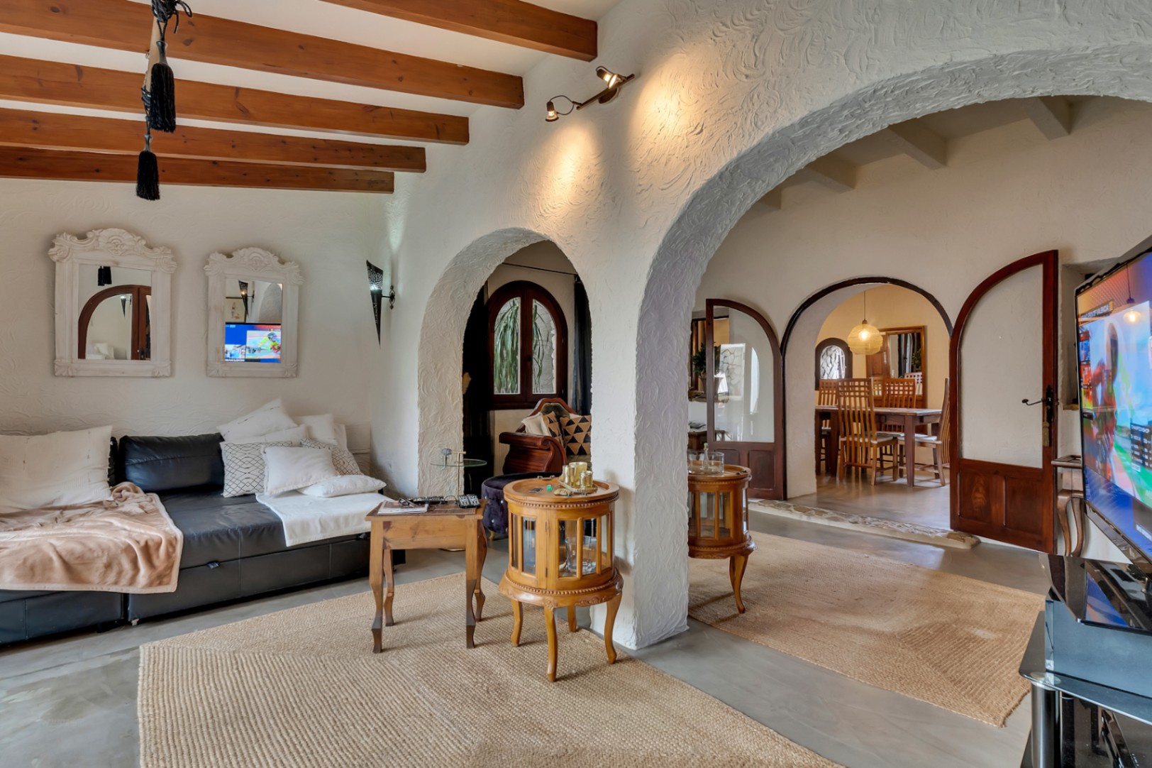 Salón con vigas de madera vistas, arcos tradicionales y paredes blancas texturizadas que conectan con el comedor mediante puertas de madera acristaladas.