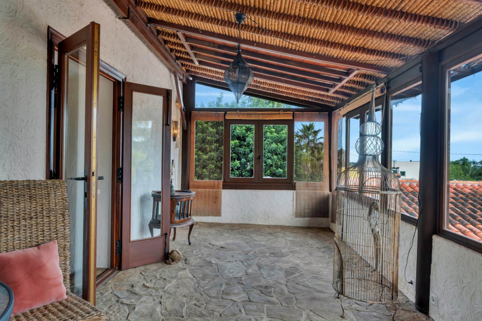 Terraza acristalada con suelo de piedra natural, techo de cañizo con vigas de madera y una lámpara de estilo marroquí, ofreciendo vistas al entorno verde.
