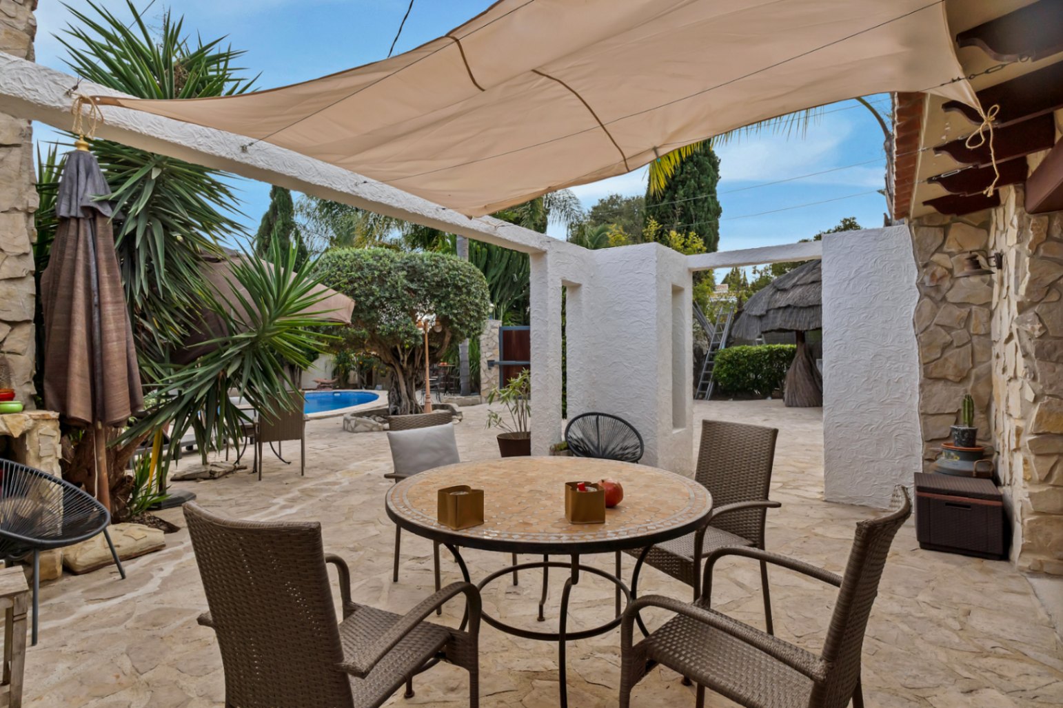 Terraza exterior con suelo de piedra natural, mesa de mosaico y toldo vela, ofreciendo vistas al jardín mediterráneo y la zona de piscina.