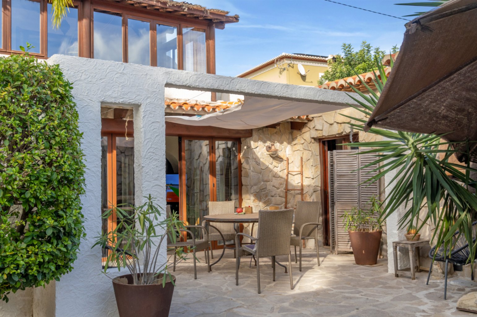 Terraza exterior con suelo de piedra natural, zona de comedor bajo toldo vela y fachada que combina piedra vista con carpintería de madera y grandes ventanales.