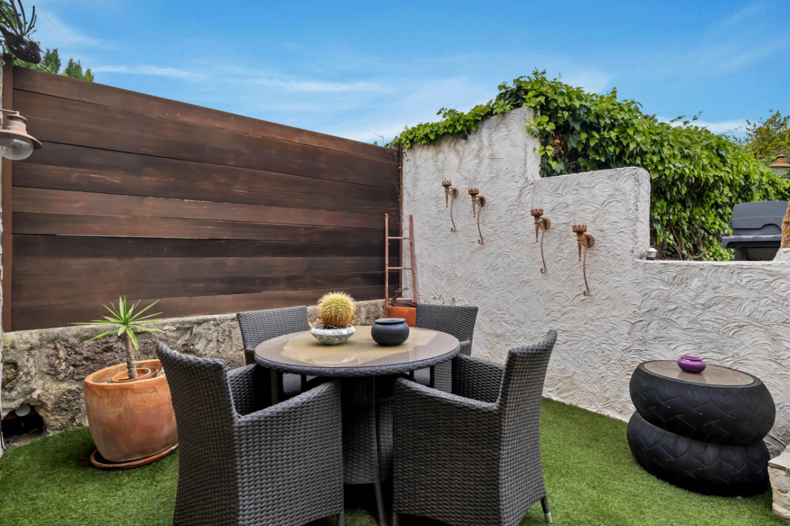 Terraza privada con césped artificial, mesa de comedor circular y sillas de ratán sintético, delimitada por muros de piedra, madera y revoco blanco.