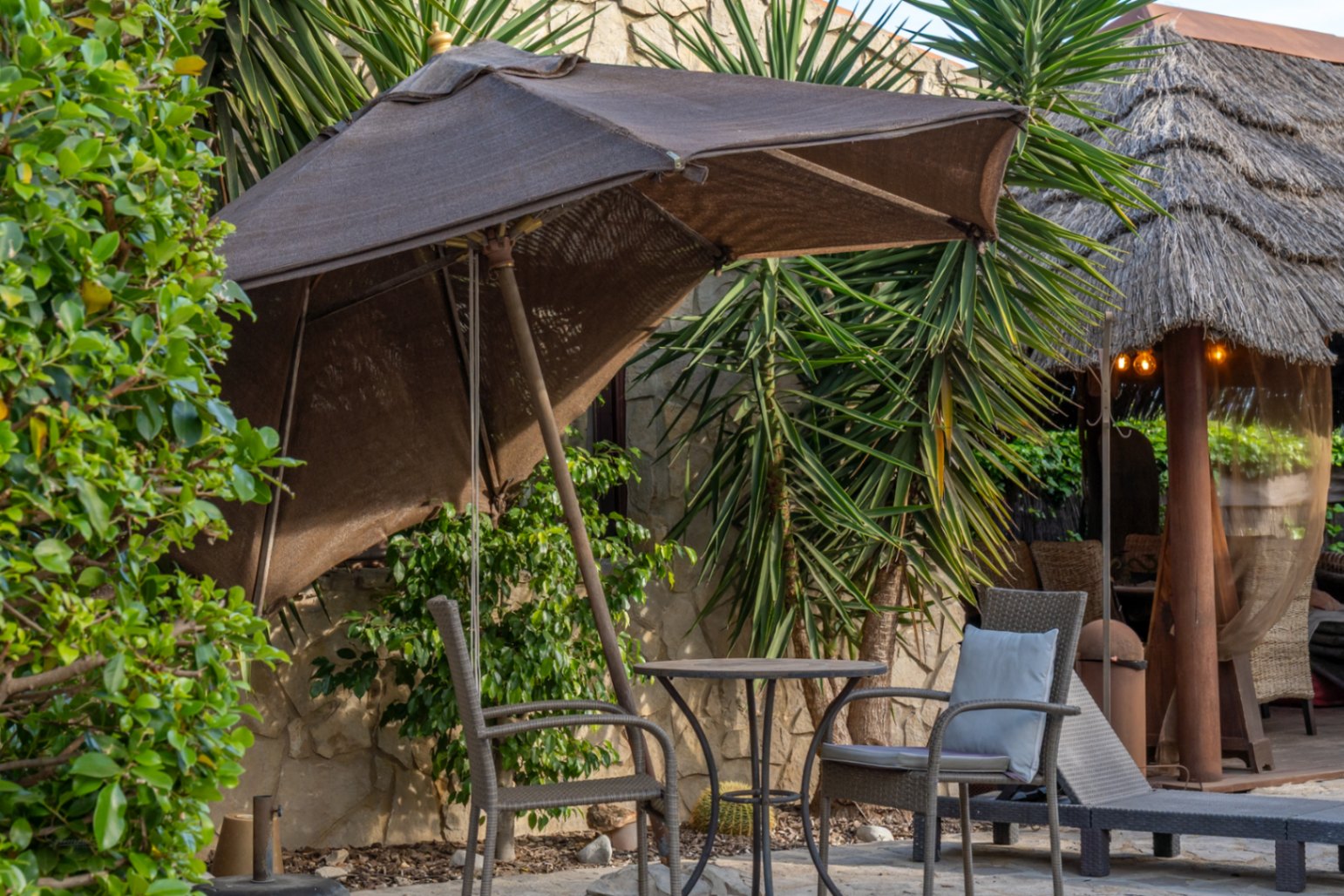 Zona de estar exterior con mobiliario de mimbre y sombrilla, situada junto a una pérgola de estilo balinés y muros de piedra natural.
