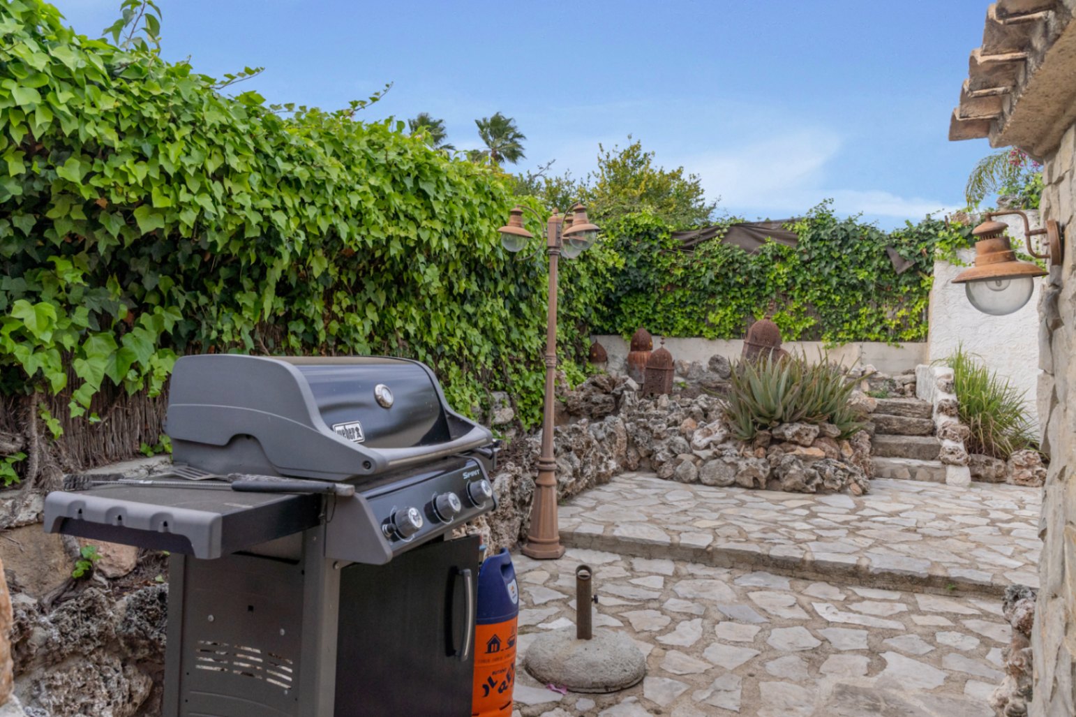 Zona exterior de barbacoa con pavimentación de piedra irregular, rodeada de muros cubiertos de hiedra y un jardín de rocalla con iluminación de estilo rústico.