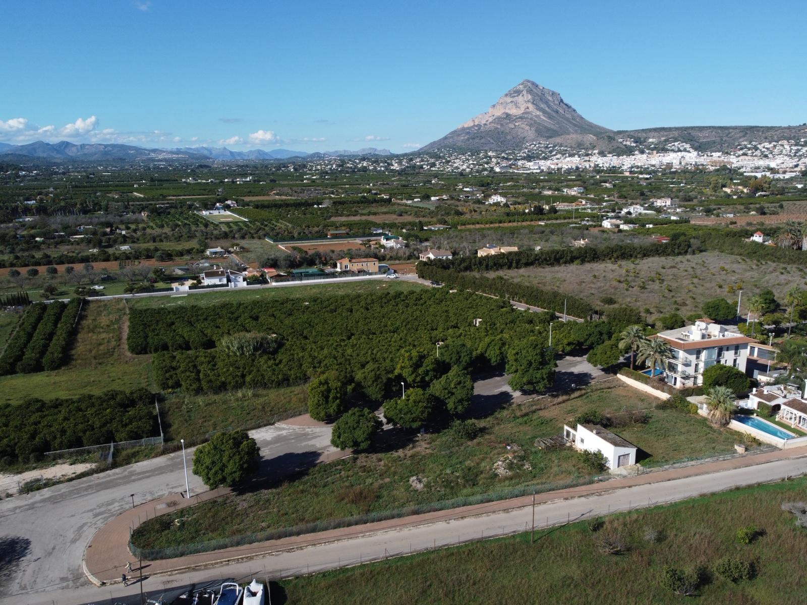 Vista aérea del solar con acceso asfaltado, rodeado de campos agrícolas y con la silueta del Montgó destacando en el horizonte bajo un cielo despejado.