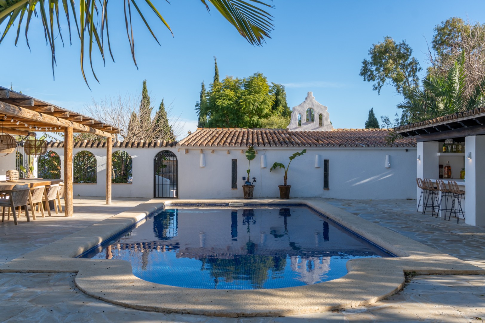 Zona exterior con piscina rodeada de pavimentos de piedra, comedor bajo pérgola de madera y barra de bar integrada en un entorno de estilo mediterráneo.