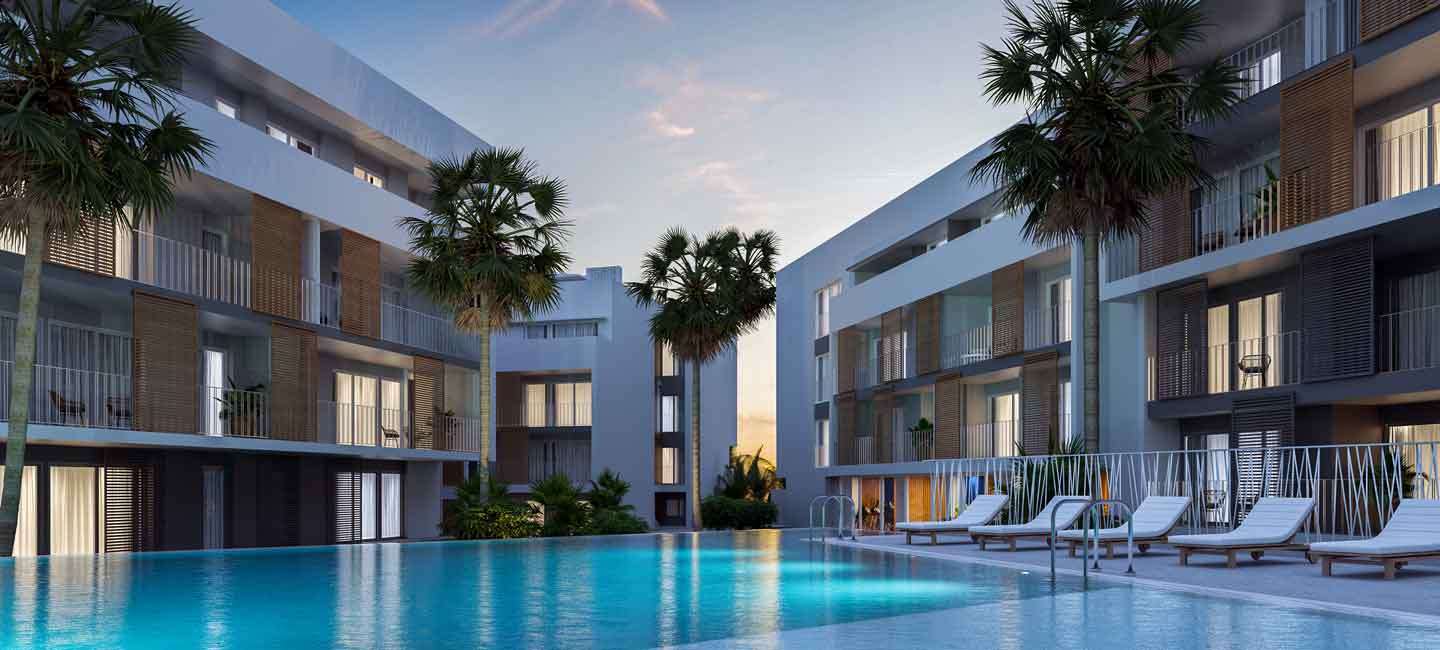 Vista exterior del complejo con piscina comunitaria, fachadas blancas acentuadas por persianas de madera y palmeras integradas bajo la luz del atardecer.