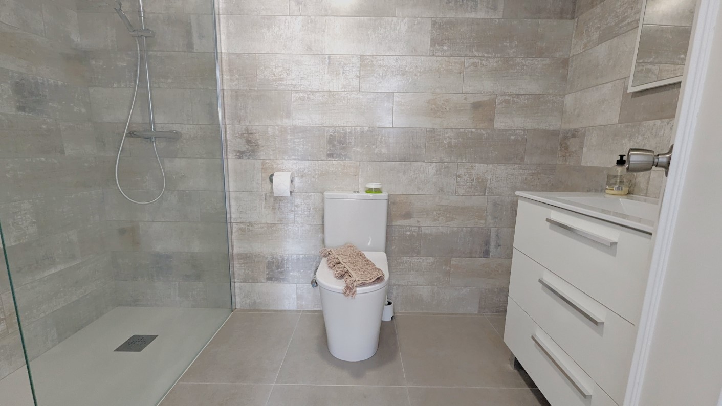 Baño moderno con ducha a ras de suelo, mampara de cristal, azulejos texturizados en tonos grises y mueble de lavabo blanco con amplios cajones.