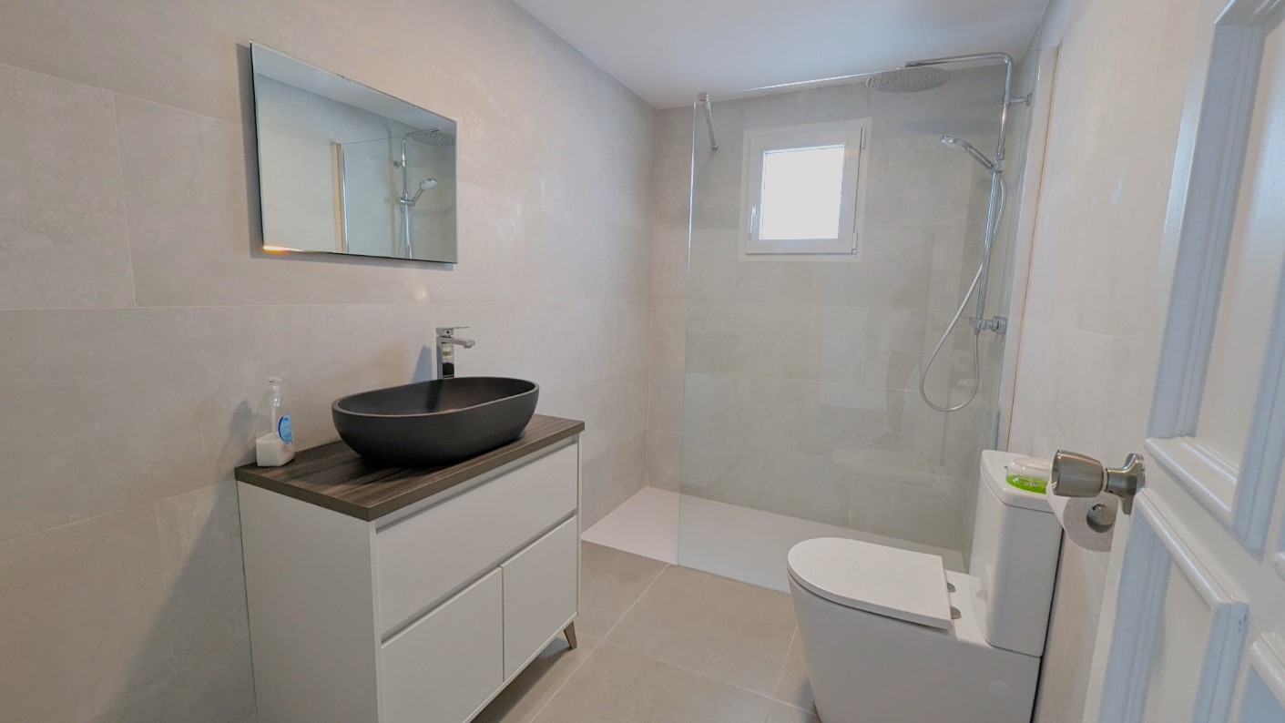 Baño moderno con lavabo de sobremesa negro, ducha a ras de suelo con mampara de cristal y revestimiento de azulejos grises de gran formato.