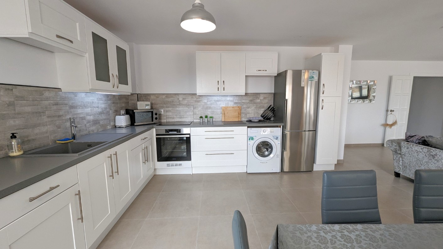 Cocina moderna de planta abierta con armarios blancos estilo shaker, salpicadero de azulejos grises efecto piedra y electrodomésticos de acero inoxidable integrados.