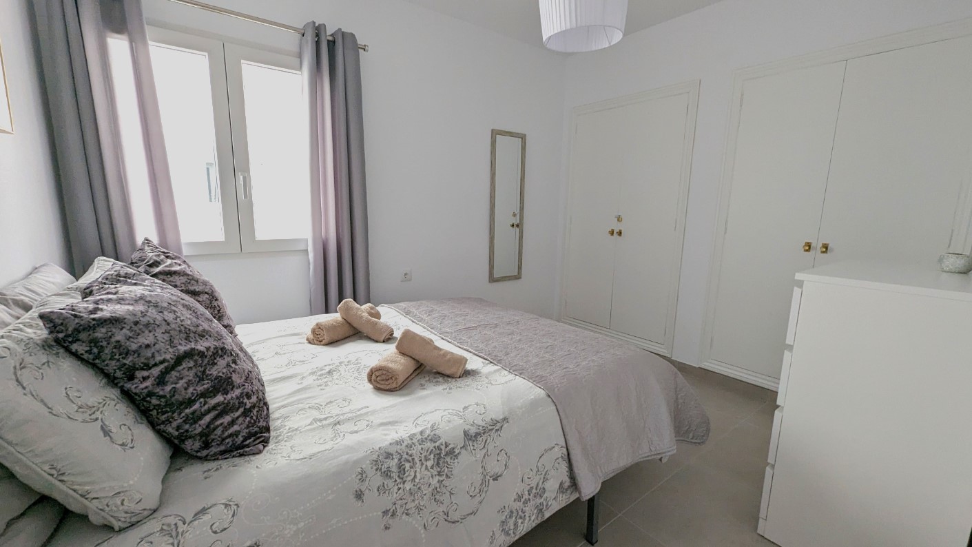 Dormitorio doble con armarios empotrados blancos, espejo de pared y suelos de baldosas. La estancia cuenta con luz natural y una decoración en tonos neutros y grises.