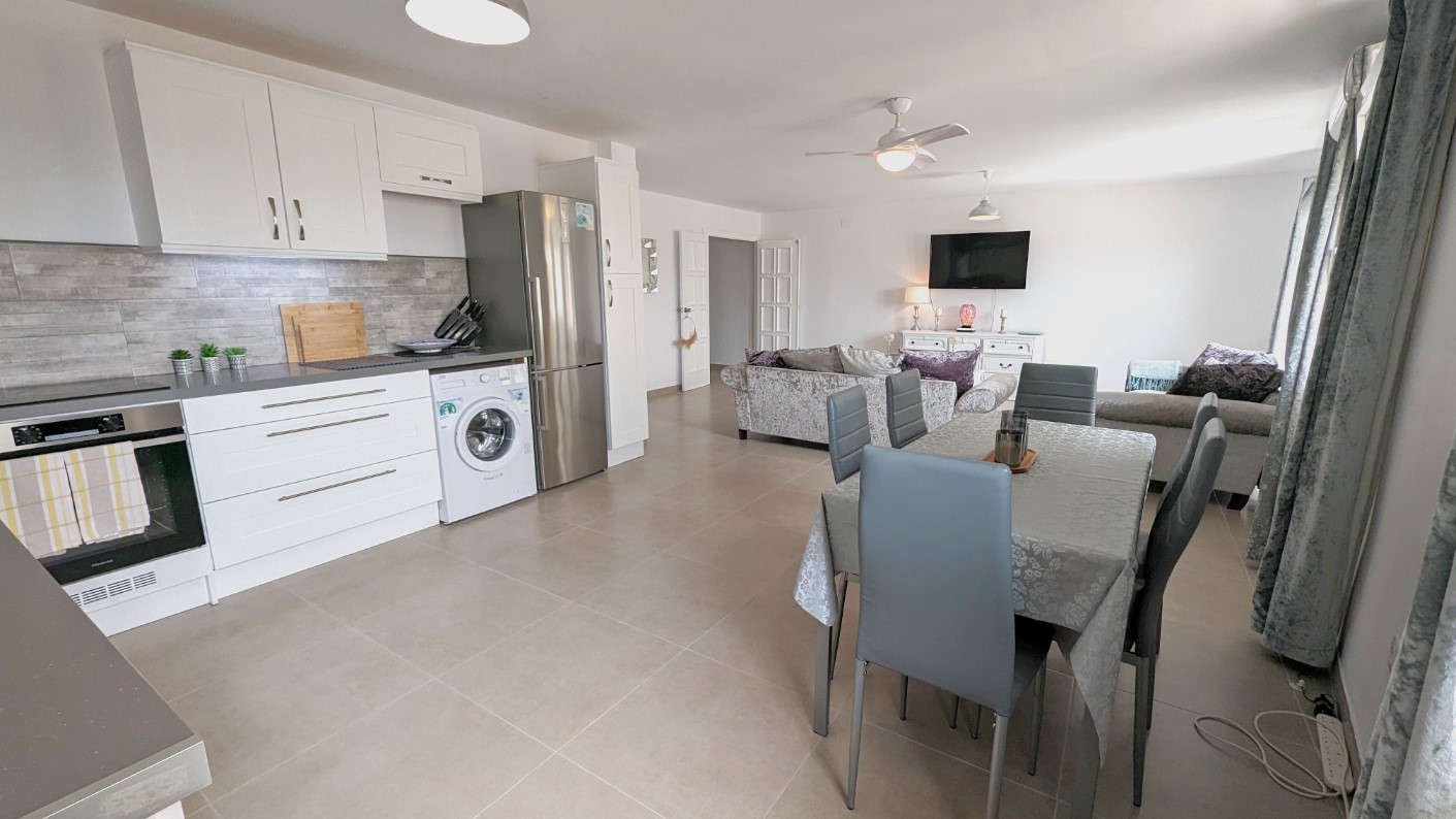 Espacio diáfano que integra una cocina blanca moderna con electrodomésticos, zona de comedor con sillas grises y salón con ventilador de techo y suelos cerámicos.