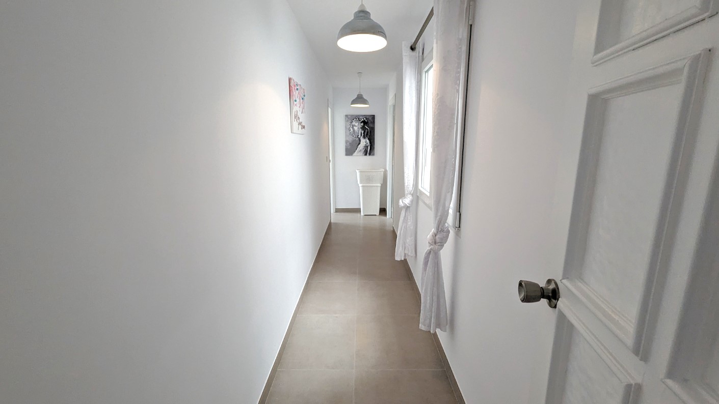 Pasillo luminoso con paredes blancas, suelos de gres, lámparas colgantes de estilo industrial y ventanas con cortinas blancas anudadas.