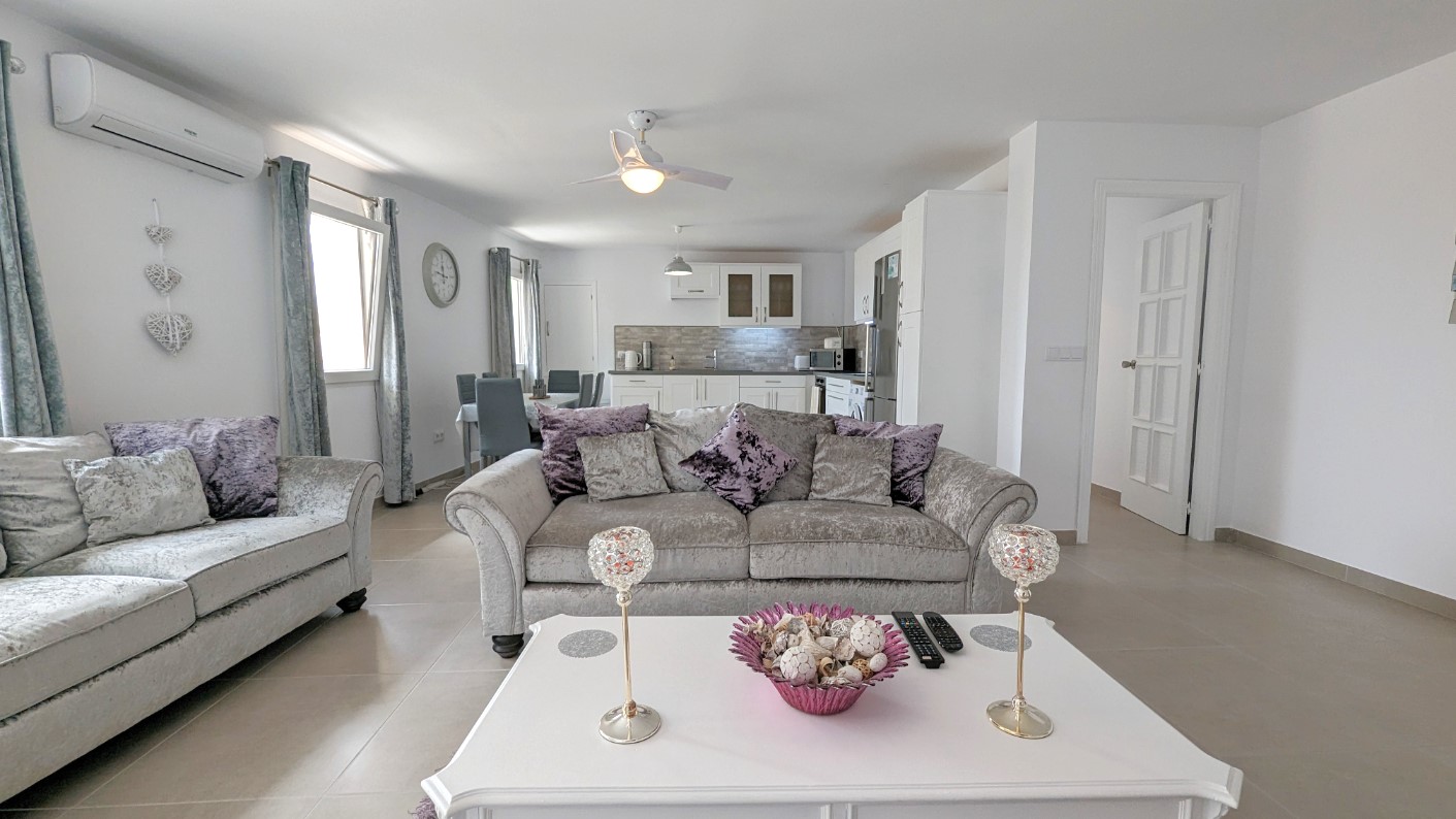 Salón de planta abierta con sofás de terciopelo gris, mesa de centro blanca y ventilador de techo. Incluye zona de comedor y cocina blanca integrada con suelos cerámicos.