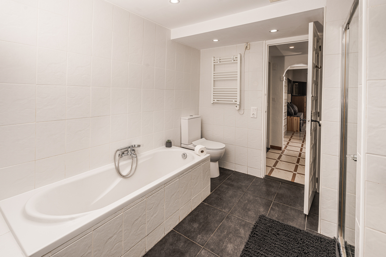 Baño funcional con bañera alicatada en blanco, suelo de gres oscuro, radiador toallero y focos empotrados en el techo.