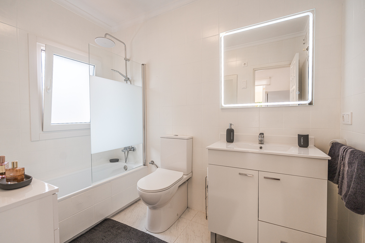 Baño moderno con revestimiento cerámico blanco, mueble lavabo con espejo de iluminación LED y bañera con mampara de cristal y ducha de efecto lluvia.