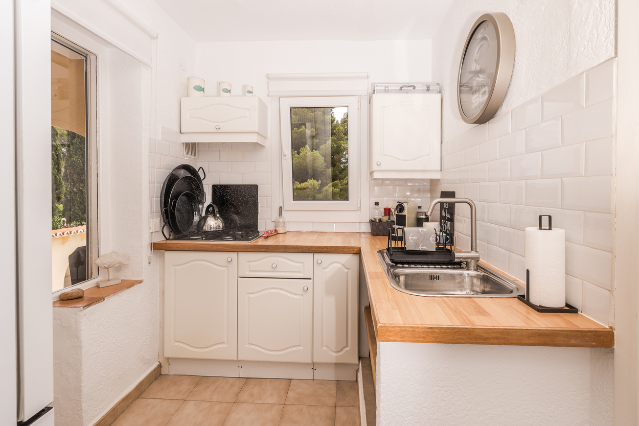 Cocina compacta con armarios blancos, encimeras de madera y salpicadero de azulejos tipo metro. Incluye encimera de gas, fregadero de acero inoxidable y vistas al jardín.