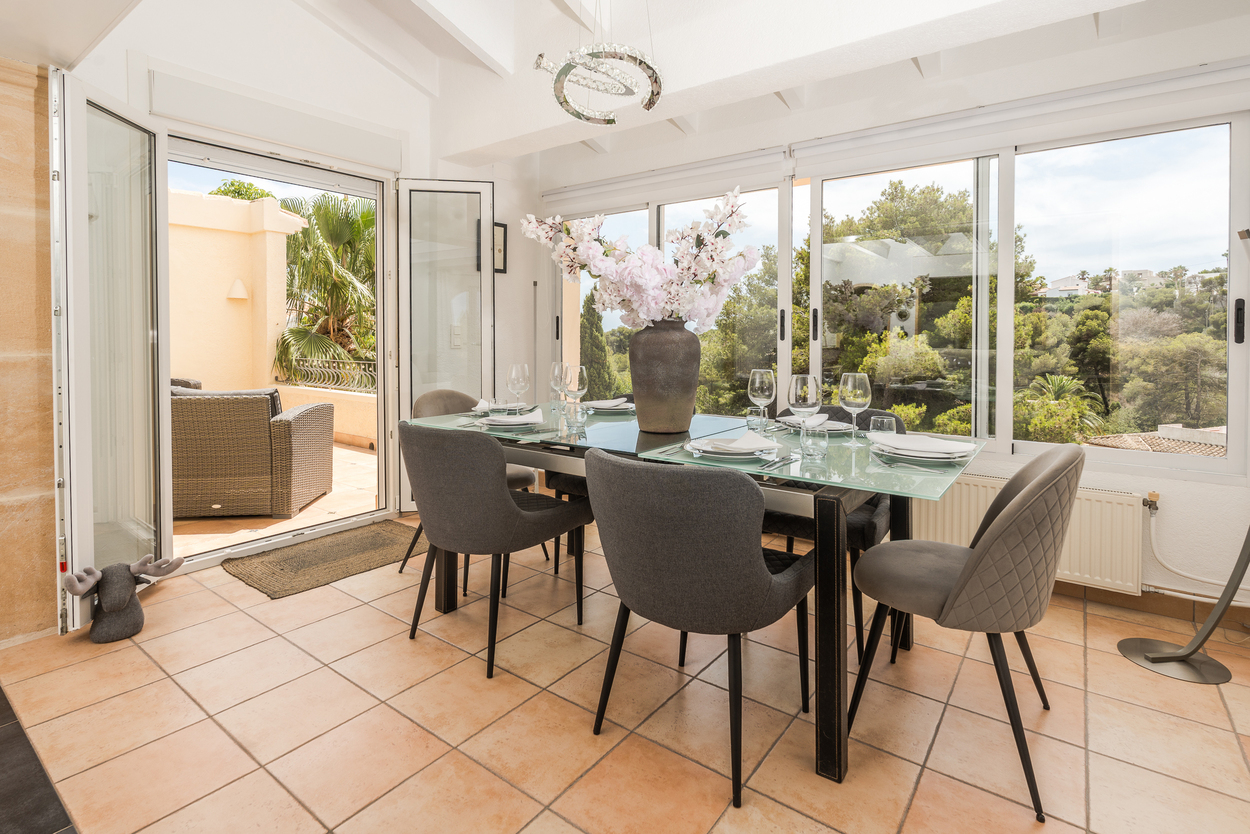 Comedor luminoso con suelos de terracota, techos de vigas blancas y amplios ventanales que conectan con la terraza y ofrecen vistas al entorno natural de Javea.
