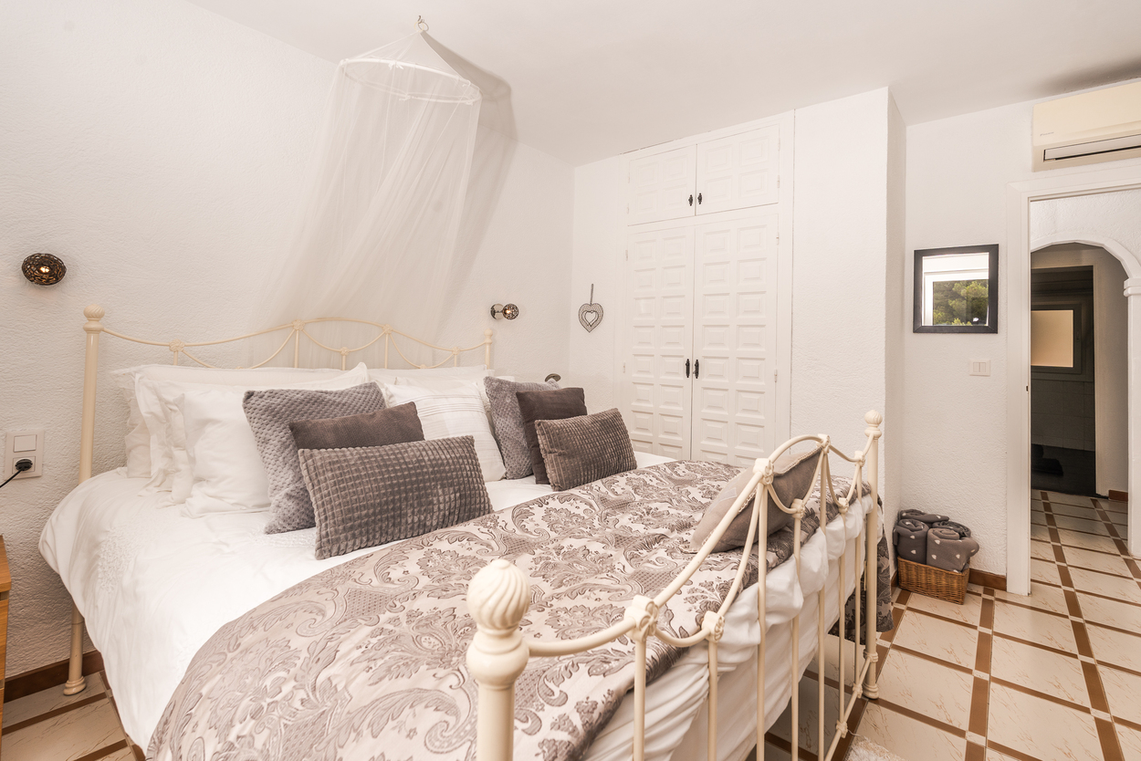 Dormitorio con cama de hierro forjado blanco, dosel de mosquitera y armarios empotrados. Incluye suelos cerámicos y aire acondicionado sobre el arco de entrada.