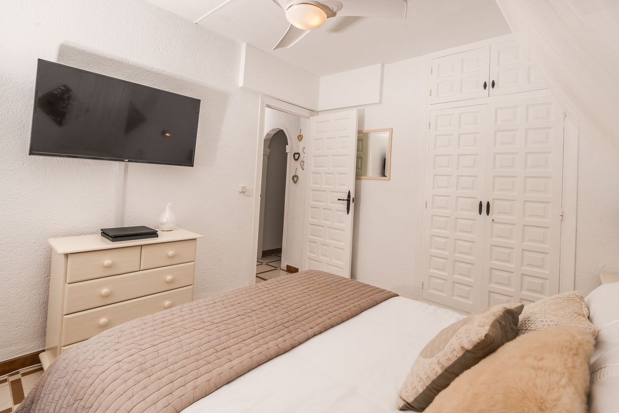 Dormitorio luminoso con armarios empotrados de paneles blancos, televisión de pared y ventilador de techo. Incluye cómoda a juego y suelos cerámicos tradicionales.