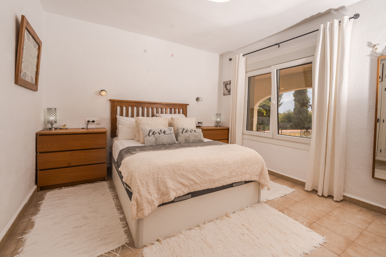 Dormitorio luminoso con cabecero de madera, ventanas de doble acristalamiento y suelo de baldosas. Incluye cómodas de madera a juego y vistas al jardín exterior.