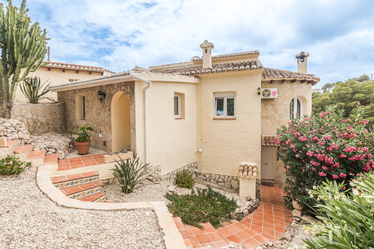 Fachada mediterránea con muros de piedra natural, escaleras de terracota decoradas y jardín de bajo mantenimiento con cactus y adelfas en flor.