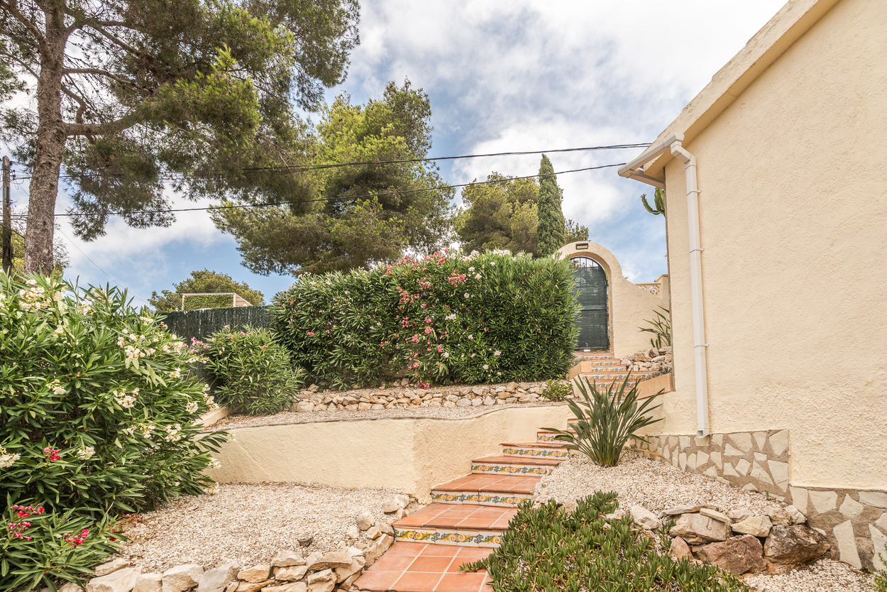 Jardín mediterráneo aterrazado con escaleras de terracota y azulejos decorativos, rodeado de adelfas y grava, que conduce a una puerta de entrada arqueada.