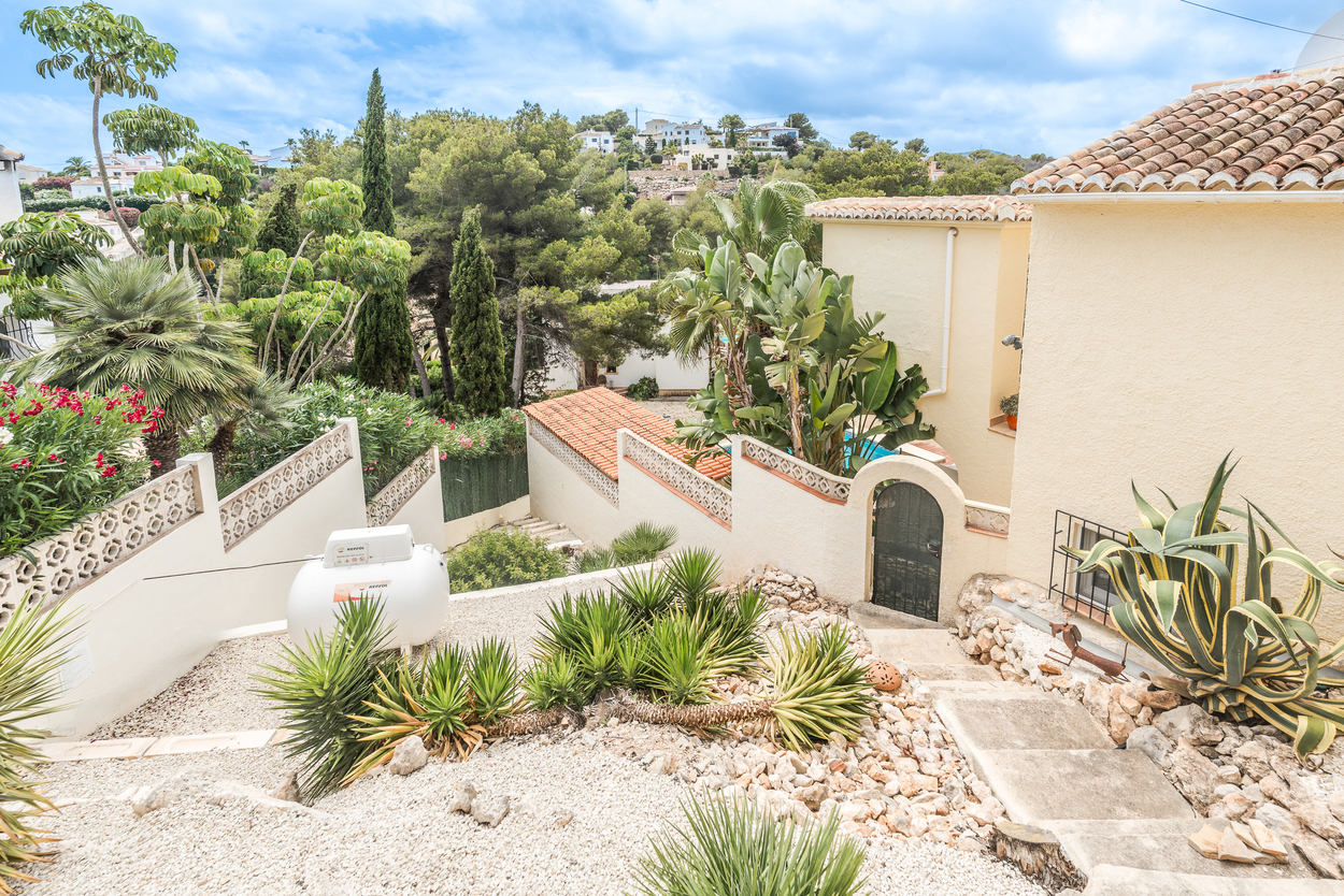 Jardín mediterráneo aterrazado con grava blanca, yucas y agaves. Escaleras de hormigón descienden hacia una entrada de arco con puerta de forja y muros encalados.