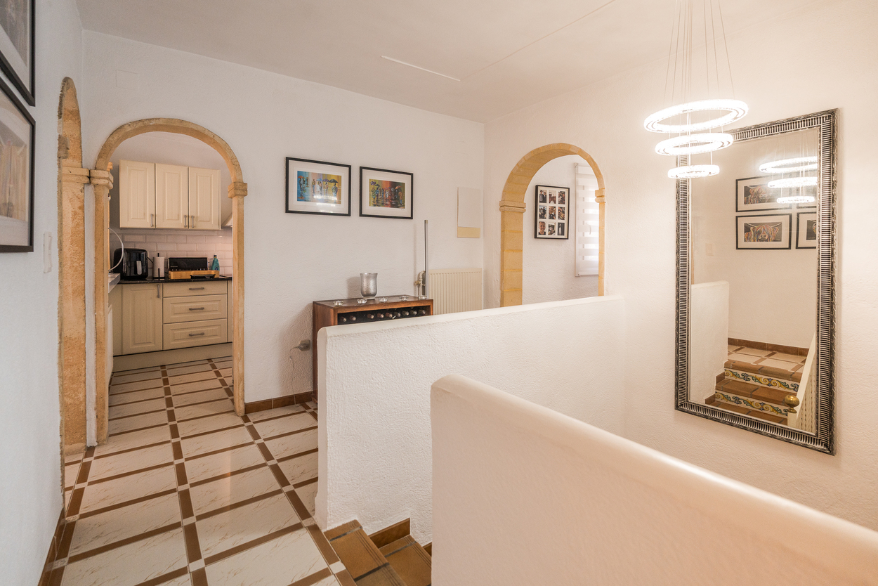 Pasillo con arcos de piedra tosca, suelos cerámicos y lámpara circular moderna. Se aprecia el acceso a la cocina y una escalera con azulejos decorativos.