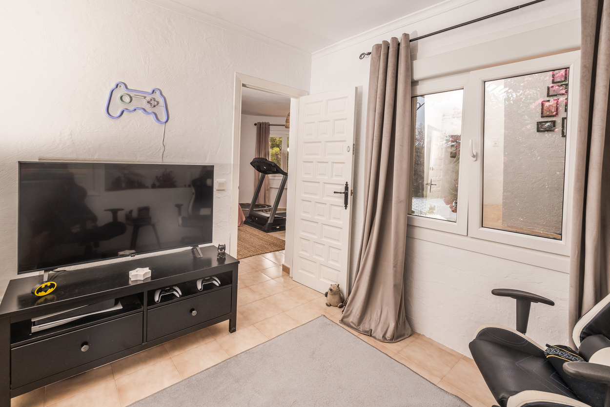 Sala multimedia con zona de juegos, televisor de pantalla plana y suelos cerámicos, con vista a un área de gimnasio contigua con cinta de correr.