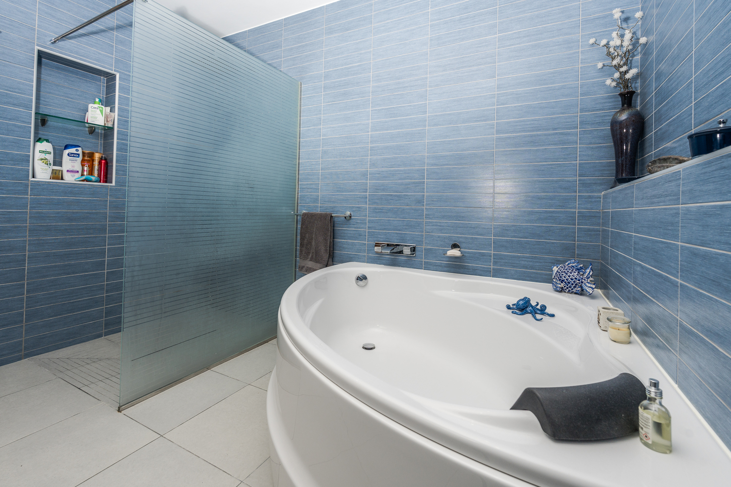 Baño moderno con bañera ovalada y ducha a ras de suelo con mampara de cristal esmerilado, revestido con azulejos azules de textura lineal y suelo gris claro.