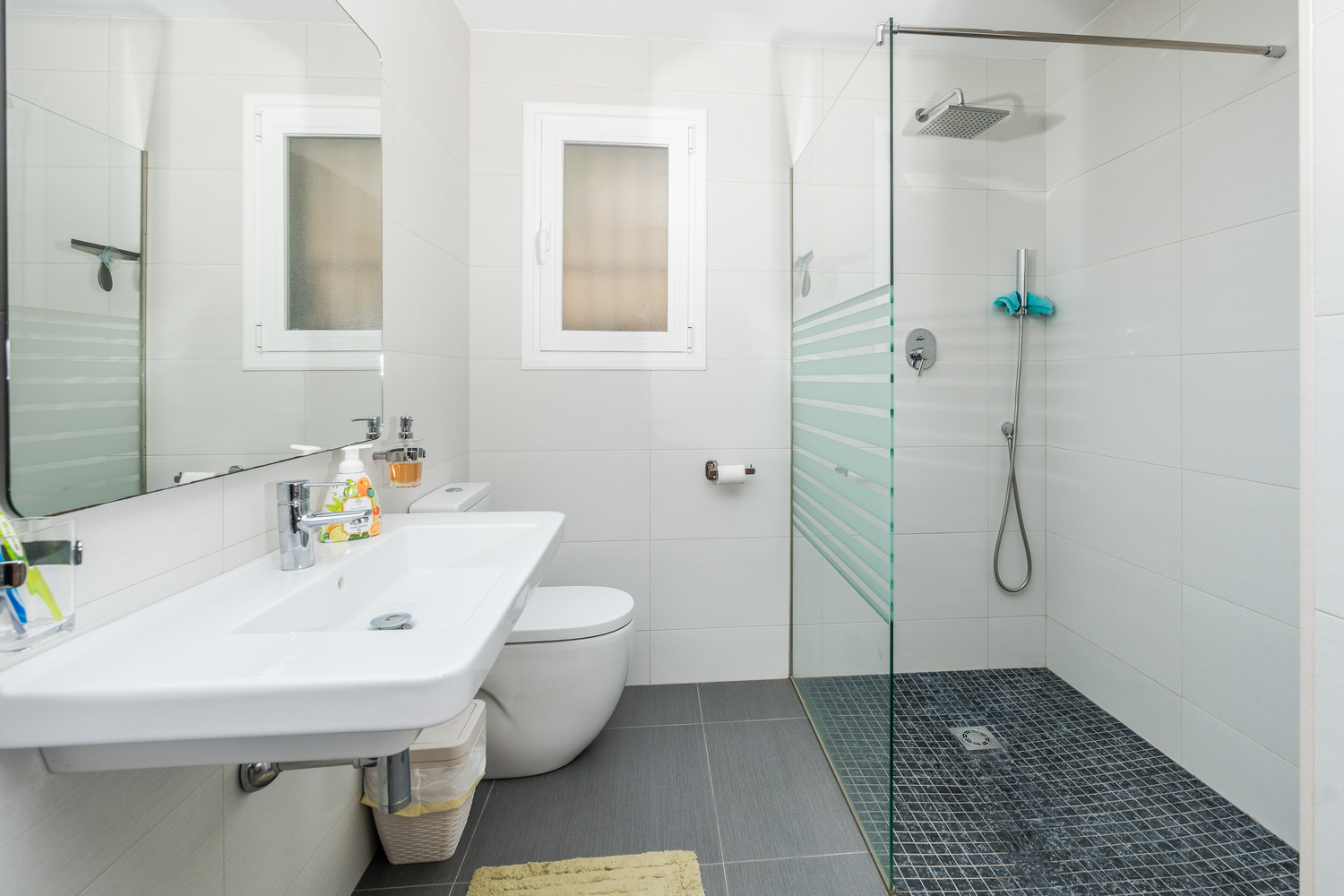 Baño moderno con ducha a ras de suelo, mampara de cristal y suelo de mosaico oscuro. Dispone de lavabo suspendido, sanitarios blancos y ventana para luz natural.