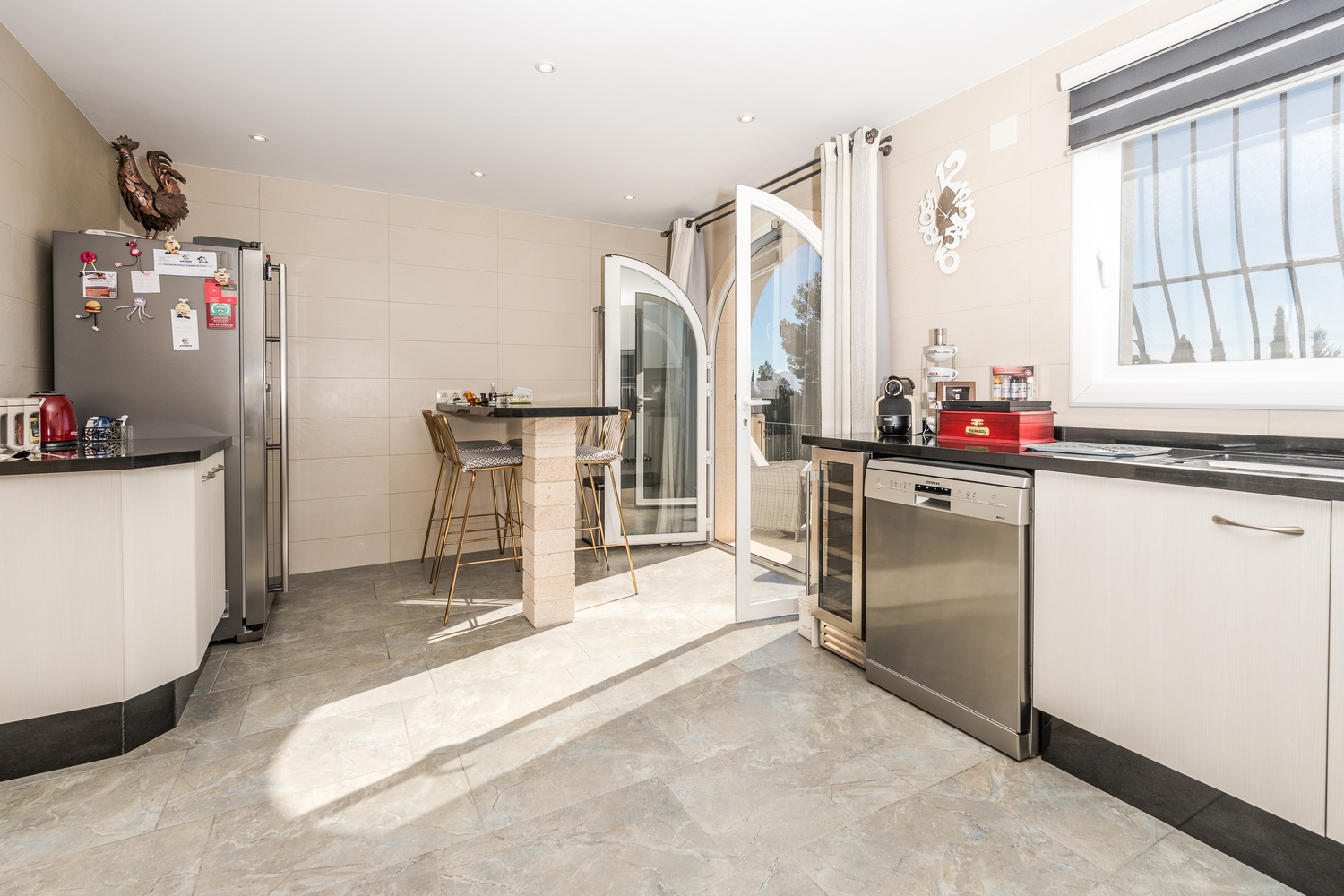 Cocina moderna con armarios blancos, encimeras de granito negro y barra de desayuno. Incluye electrodomésticos de acero inoxidable y acceso a la terraza por una puerta arqueada.