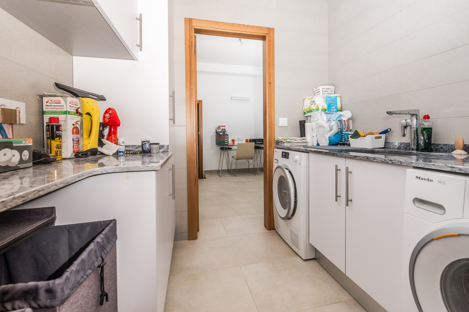Cuarto de lavandería funcional con armarios blancos, encimeras de mármol gris y electrodomésticos Miele, conectado visualmente con una zona de despacho.
