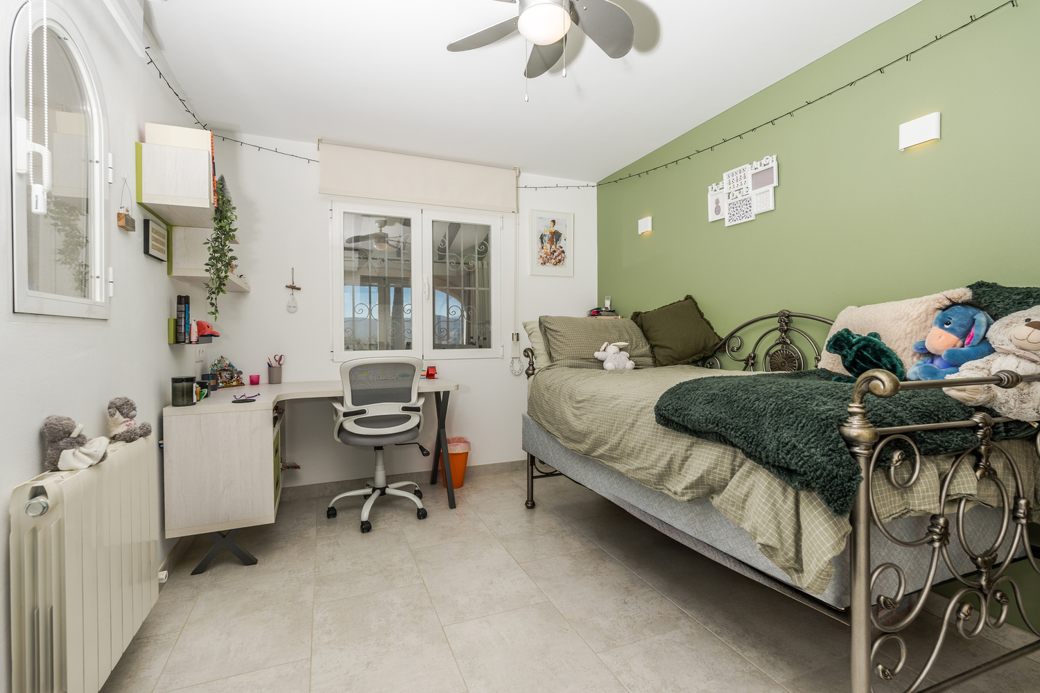 Dormitorio con pared de acento verde, cama de forja ornamental y zona de trabajo integrada con escritorio y silla ergonómica. Incluye ventilador de techo y radiador.
