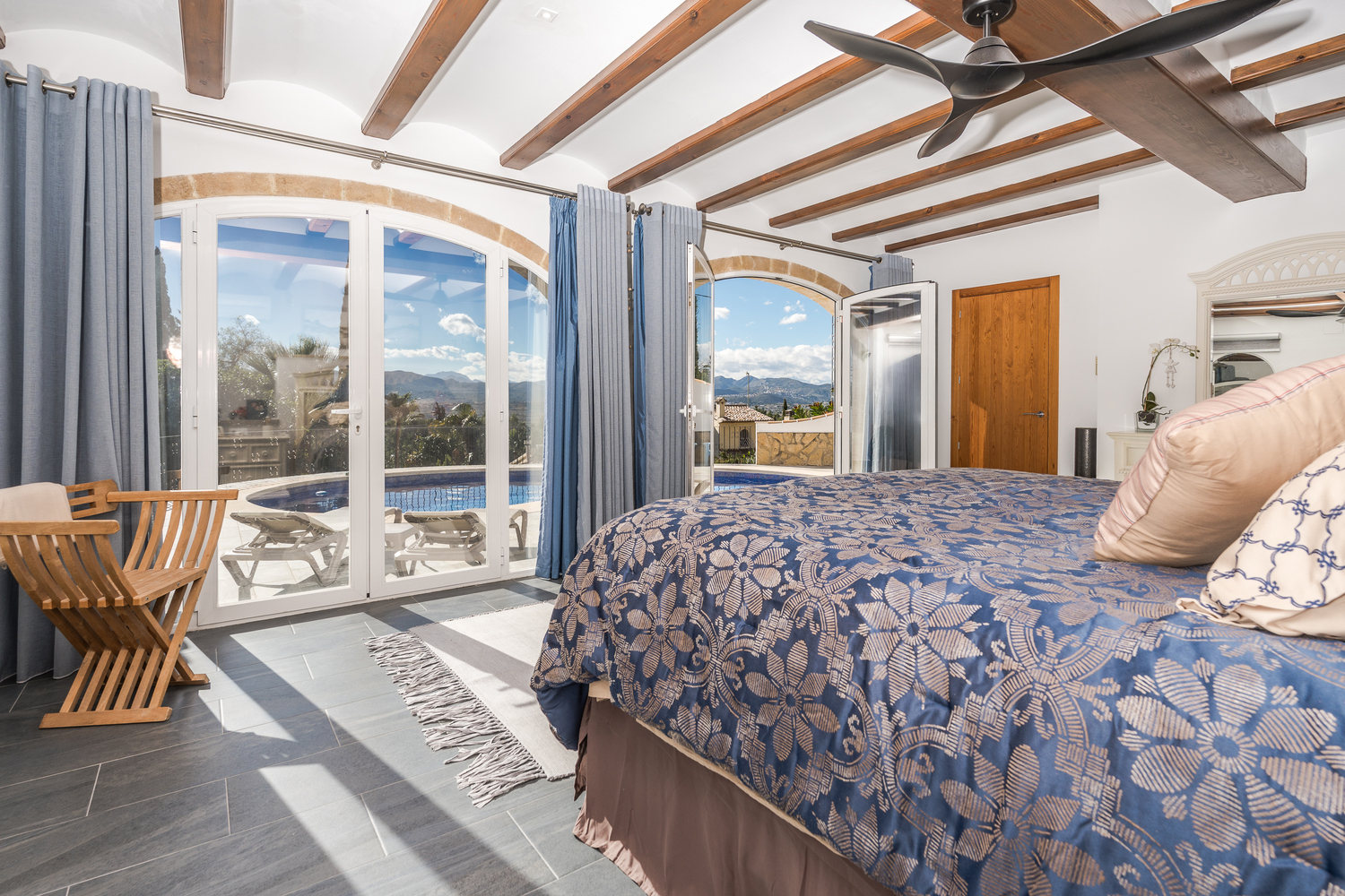 Dormitorio principal con vigas de madera, arcos de piedra natural y puertas acristaladas que conectan directamente con la zona de la piscina.