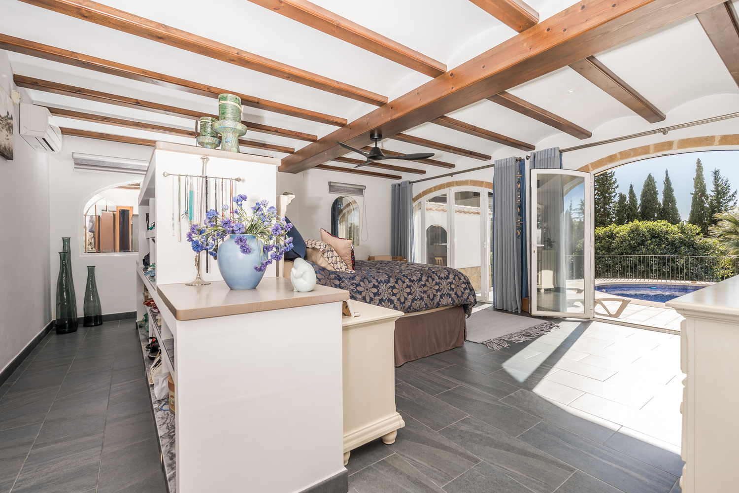 Dormitorio principal con vigas de madera vista, suelos de gres oscuro y puertas arqueadas de cristal que ofrecen acceso directo a la zona de la piscina.