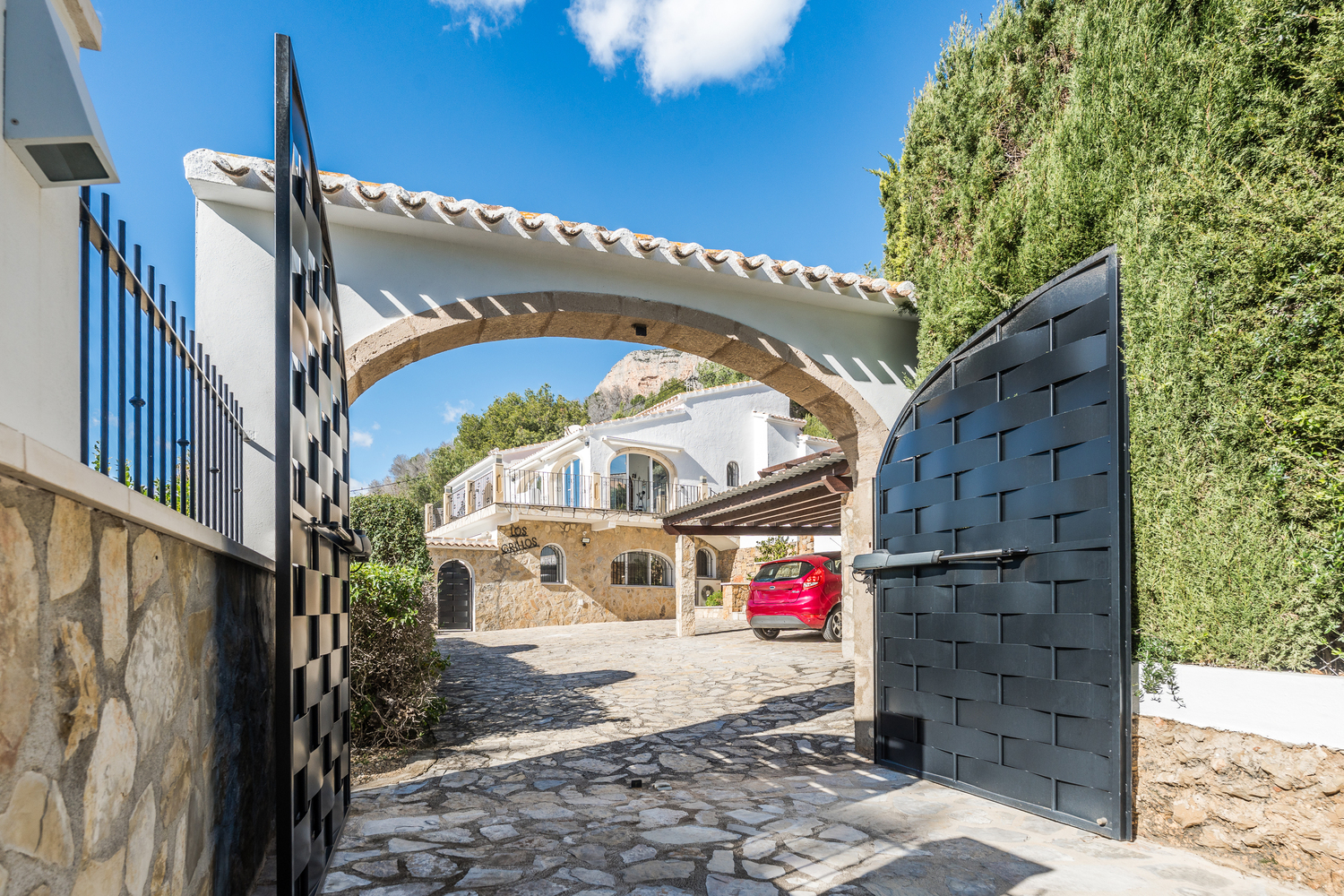 Entrada con puertas automáticas de metal negro y un arco de piedra Tosca, que da paso a un camino empedrado con vistas a la fachada de la villa y el aparcamiento.