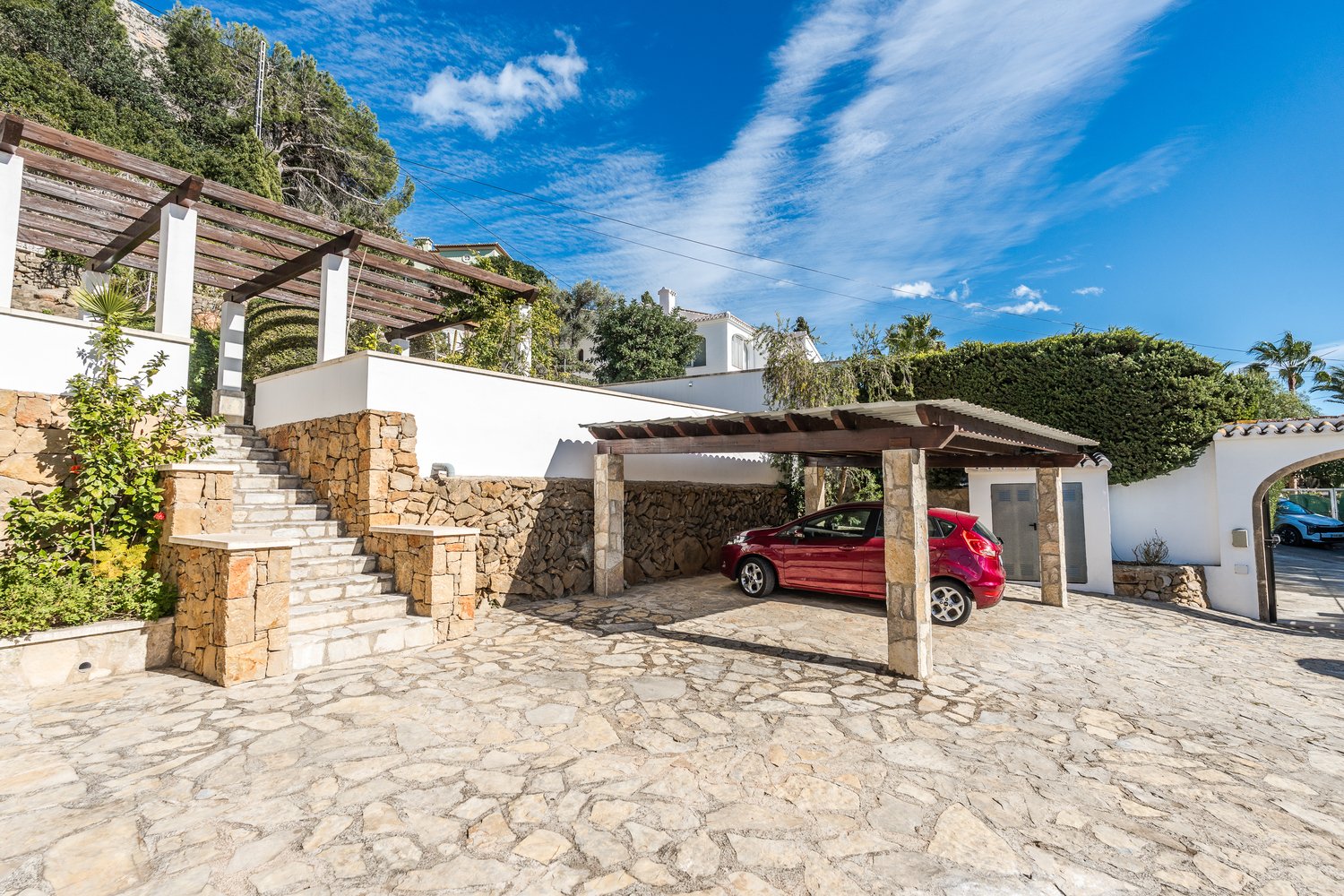 Entrada exterior con suelo de piedra irregular, un porche cubierto para vehículos y escaleras de piedra natural que conectan con los niveles superiores.