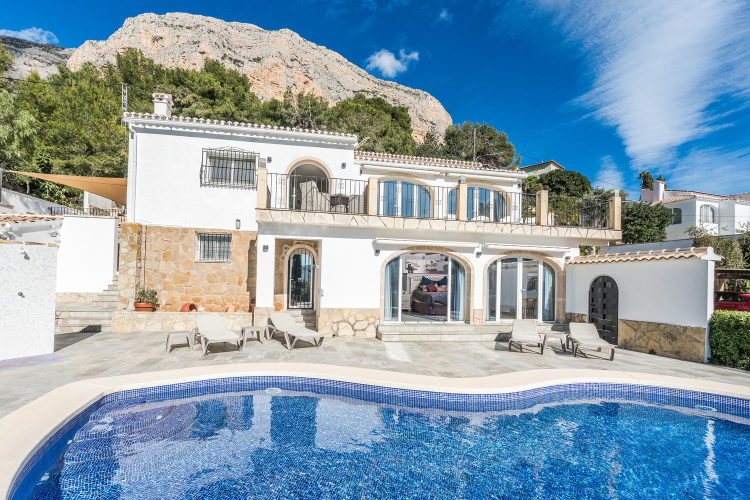 Exterior de la villa con piscina de gresite azul, terraza de piedra con tumbonas y arquitectura tradicional bajo el macizo del Montgó.