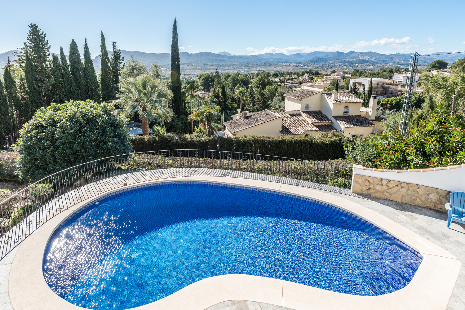 Piscina en forma de riñón con azulejos de mosaico azul, rodeada de una terraza de piedra y barandilla de hierro, con vistas panorámicas al valle y las montañas.