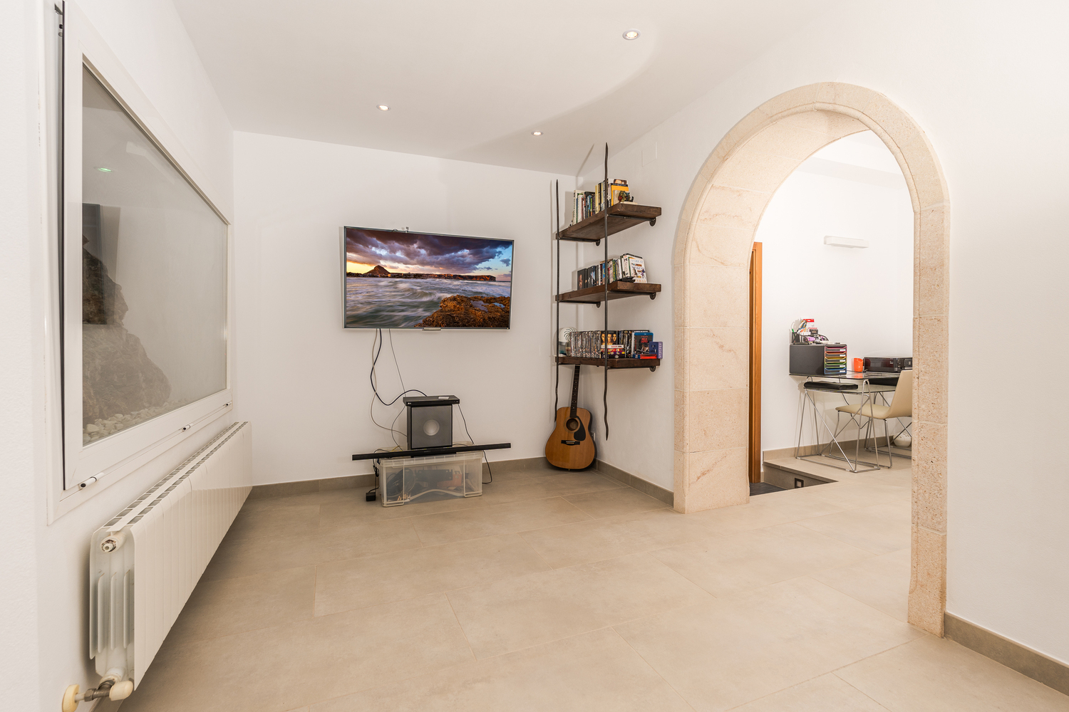 Sala de TV con pantalla de pared, estanterías de madera rústica y un arco de piedra natural que comunica con una zona de despacho independiente.