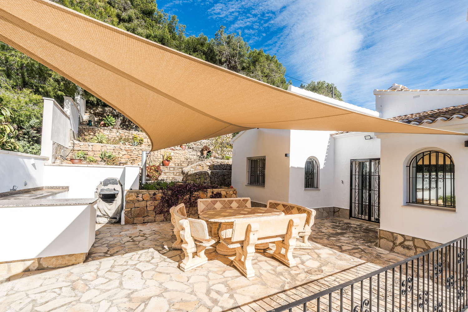 Terraza comedor exterior con mobiliario de piedra tallada bajo un toldo vela, rodeada de muros de piedra natural y vegetación mediterránea autóctona.