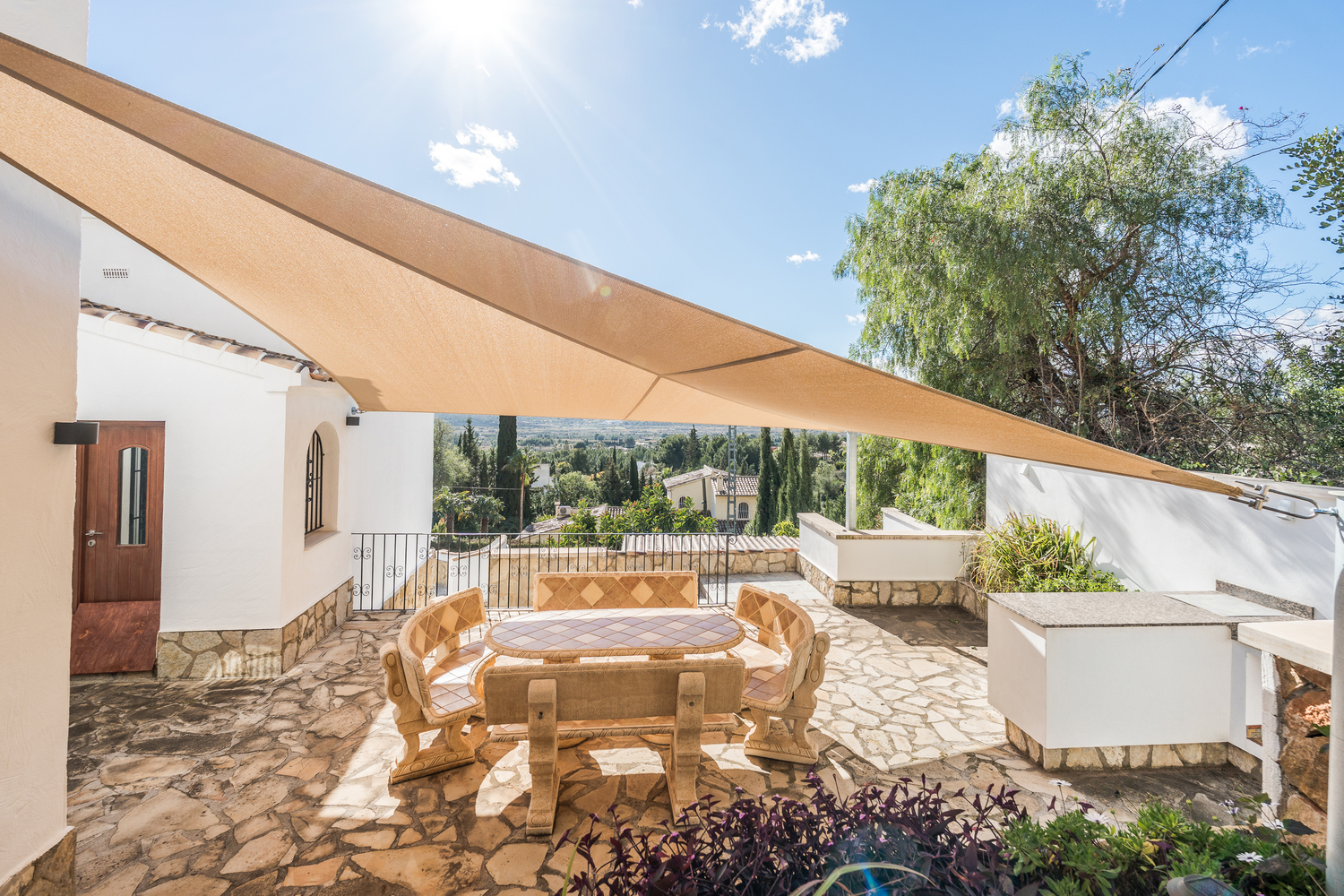 Terraza exterior con mobiliario de piedra bajo un toldo vela, que ofrece vistas panorámicas al valle y cuenta con una zona de cocina de verano.