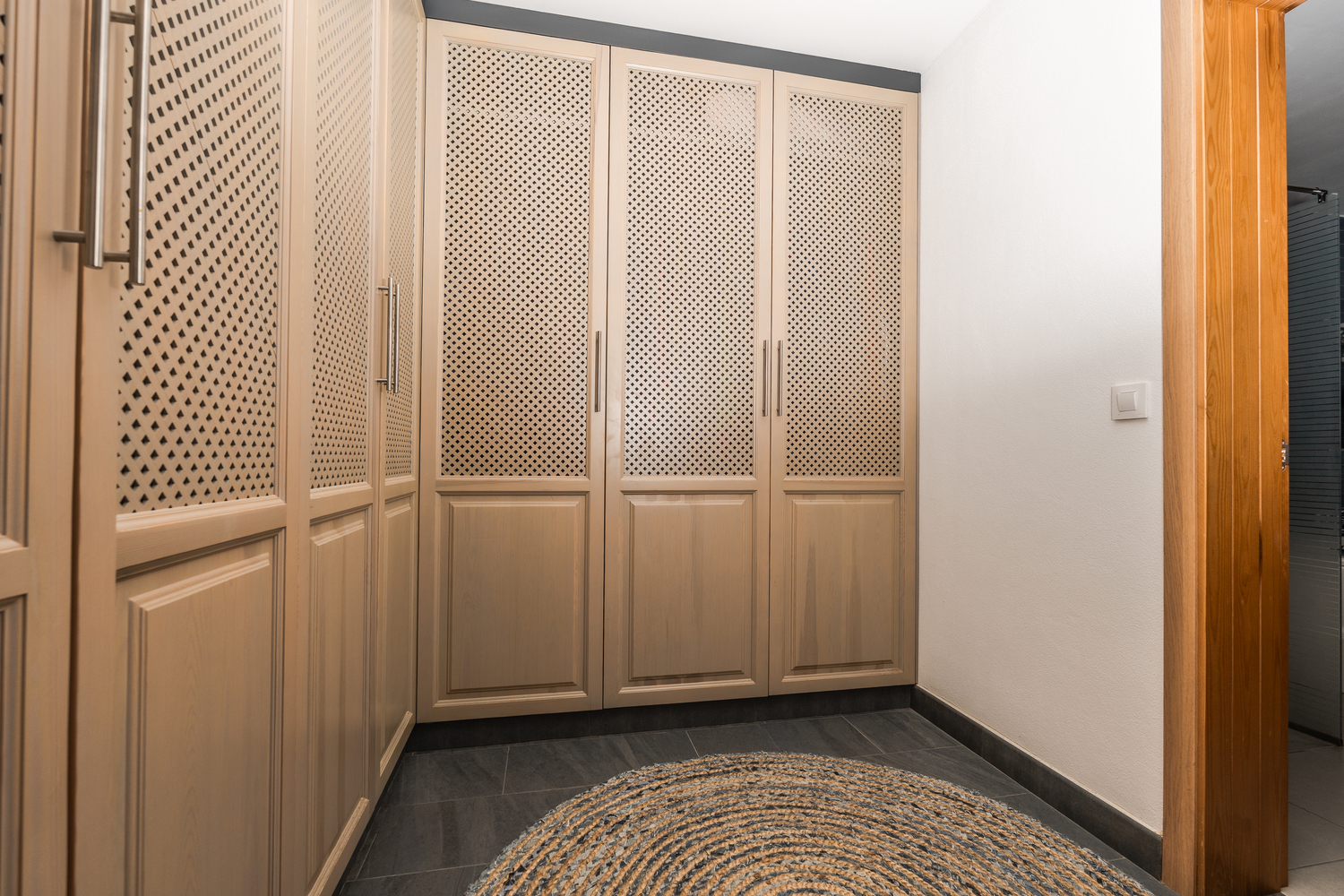 Vestidor con armarios empotrados de madera clara y paneles de celosía, suelo de baldosas oscuras y una alfombra circular de yute.
